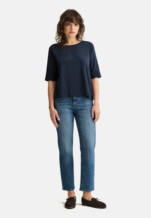 Kvinde med krøllet hår iført en løs mørkeblå top, lige blå jeans og mørke mokkasinsko, stående mod en ensfarvet baggrund.