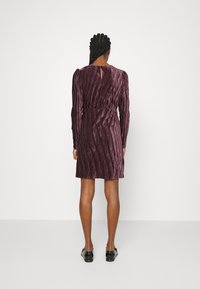 Mujer de pie de espaldas, vistiendo un vestido largo de mangas largas con pliegues de terciopelo púrpura y mocasines negros, contra un fondo blanco liso.