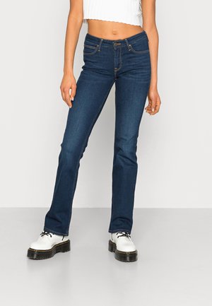 Femme portant un jean slim bleu foncé, un top court blanc côtelé, et des chaussures blanches à plateforme à lacets, debout sur un fond uni.