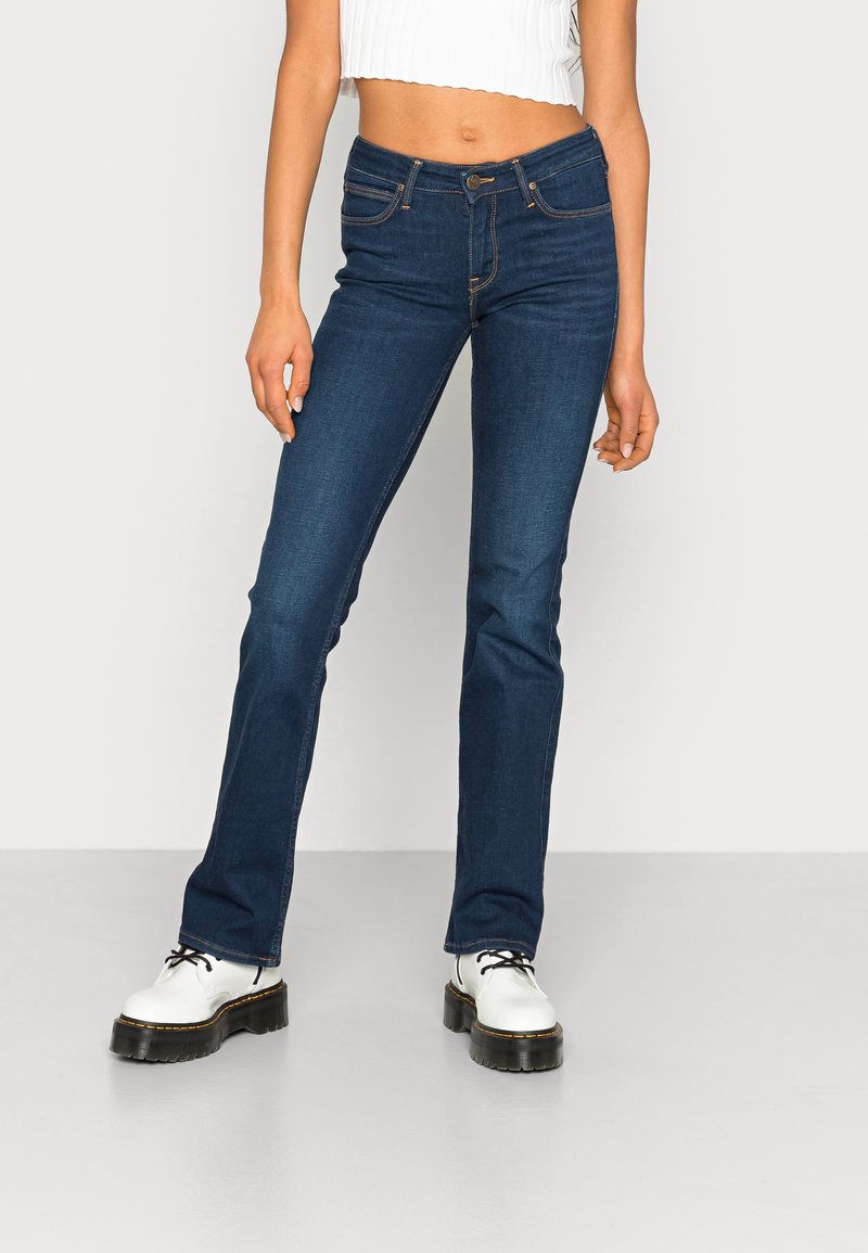 Femme portant un jean slim bleu foncé, un top court blanc côtelé, et des chaussures blanches à plateforme à lacets, debout sur un fond uni.