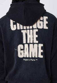 Sudadera navy con un gran texto blanco desgastado "CAMBIA EL JUEGO" en la parte posterior, con capucha ajustable y textura de tela suave.