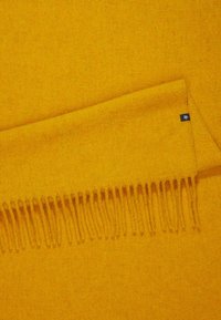 Couverture en laine jaune avec une texture douce, présentant des bords frangés et une petite étiquette logo dans un coin.