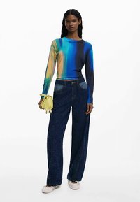 Daugiaspalvis ilgomis rankovėmis viršus su gradientiniu raštu; aukšto liemens plačios mėlynos džinsinės kelnės su žėrinčia tekstūra; laiko mažą geltoną rankinę.