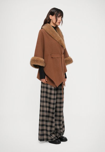Mujer con un abrigo marrón con cinturón y cuello y puños de piel, mangas largas negras, pantalones de cuadros anchos y zapatos de plataforma negros, de pie de lado.