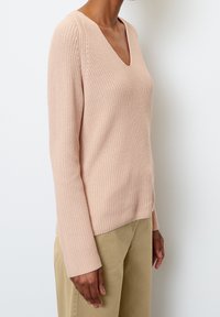 Pull rose clair en maille à col en V avec manches longues et texture côtelée, associé à un pantalon beige. Aucun motif ou accent visible.