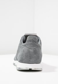 Sneaker en daim gris avec semelle blanche, vue depuis l'arrière, présentant un logo en relief subtil sur la languette arrière.