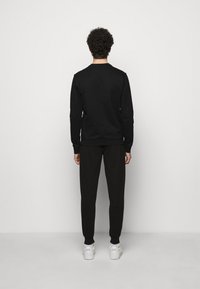 Man medLockiga hår som står vänd bort, iförd en svart sweatshirt, svarta byxor och vita EA7-sneakers mot en enkel ljus bakgrund.