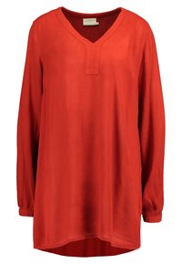 Blouse rouge à manches longues en tissu texturé avec un décolleté en V et un ourlet légèrement asymétrique. Dispose d'un patch au niveau du col.