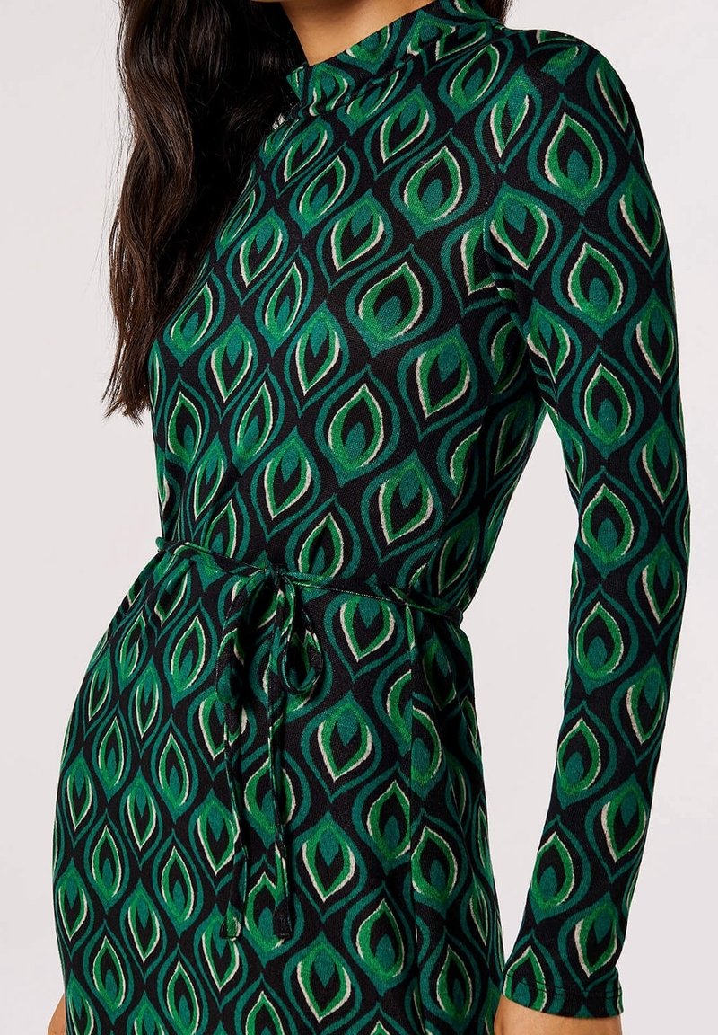 Femme portant une robe ajustée à manches longues avec des motifs de feuilles vertes et noires et une ceinture nouée à la taille.