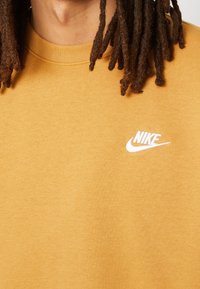 Keltainen Nike-huppari, jossa on pyöreä pääntie, pehmeä materiaali ja valkoinen logo vasemmassa rintapuoliskossa. Lähikuva.