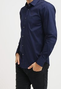 Man med en åtsittande marinblå skjorta med knappar och svarta jeans, med båda händerna i fickorna mot en enkel vit bakgrund.