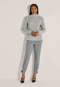 Frau trägt einen hellgrauen, gerippten Rollkragenpullover, graue, taillierte Hose mit Schlitz an den Knöcheln und schwarze spitze Absatzschuhe, steht vor einer schlichten Wand.