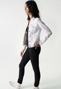 Chaqueta de mezclilla blanca con mangas largas, bolsillos delanteros y herrajes plateados, combinada con jeans ajustados negros y zapatos blancos.