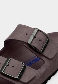 Κοντινό πλάνο καφέ σουέντ σανδαλιού Birkenstock με δύο ρυθμιζόμενες μεταλλικές αγκράφες και ορατό μπλε λογότυπο στο πέλμα.