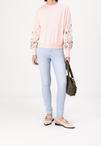 Felpa rosa chiaro con ricamo floreale sulle maniche a sbuffo, abbinata a jeans skinny azzurri e ballerine beige. Borsa verde piccola in mano.