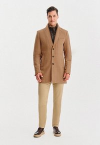 Karmelowy płaszcz wełniany z wycięciem na kołnierzu, czterema guzikami z przodu i kieszeniami, noszony na czarną kurtkę wewnętrzną, beżowe spodnie i ciemne buty.