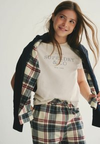 Béžové tričko s potlačou "SUPERDRY & CO", kombinované s károvanými nohavicami v krémovej, námorníckej a burgundskej farbe. Tmavý sveter prehodený cez ramená.