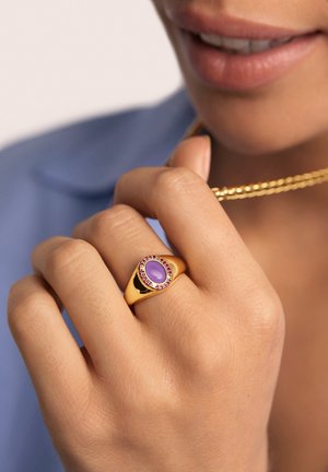Anillo de oro con una piedra ovalada morada rodeada de detalles rosados. La mano sostiene una cadena de oro sobre un fondo azul claro.