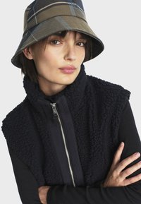 Čierny fleece vestový sveter so zipsom, preložený cez čierny top. Oblečenie dopĺňa béžová kockovaná bucket čiapka.