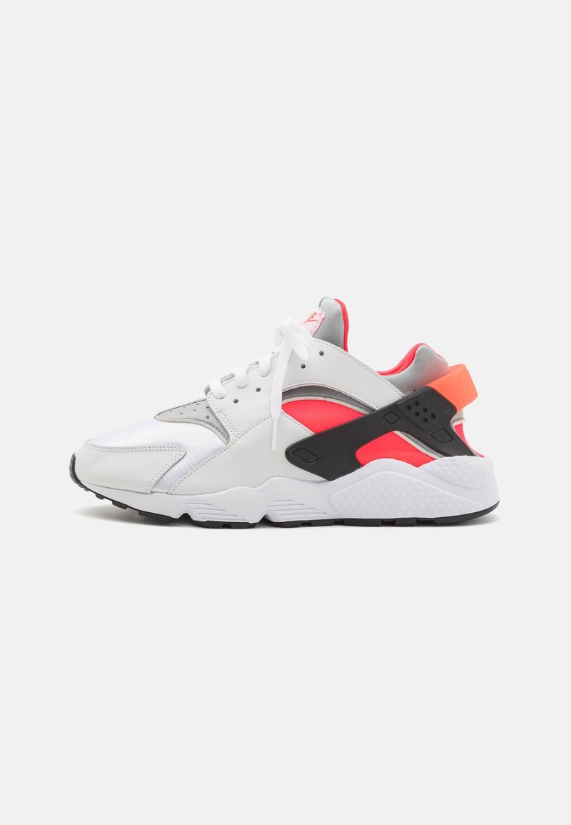 Baskets de sport blanches, grises et orange néon avec une bride de talon noire et des lacets blancs, vues de profil sur un fond clair uni.