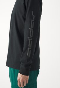 Black long-sleeve shirt made of cotton, featuring a grey logo printed vertically along the right sleeve. Paired with green textured pants.