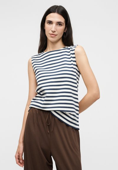 Femme aux longs cheveux foncés portant un haut sans manches blanc rayé bleu marine et un pantalon marron ample, se tenant devant un fond uni.
