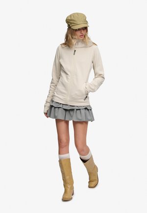 Kvinde iført beige jakke, lysegrå flæse-nederdel, brune støvler, hvide sokker og beige kasket, står med den ene hånd i lommen.