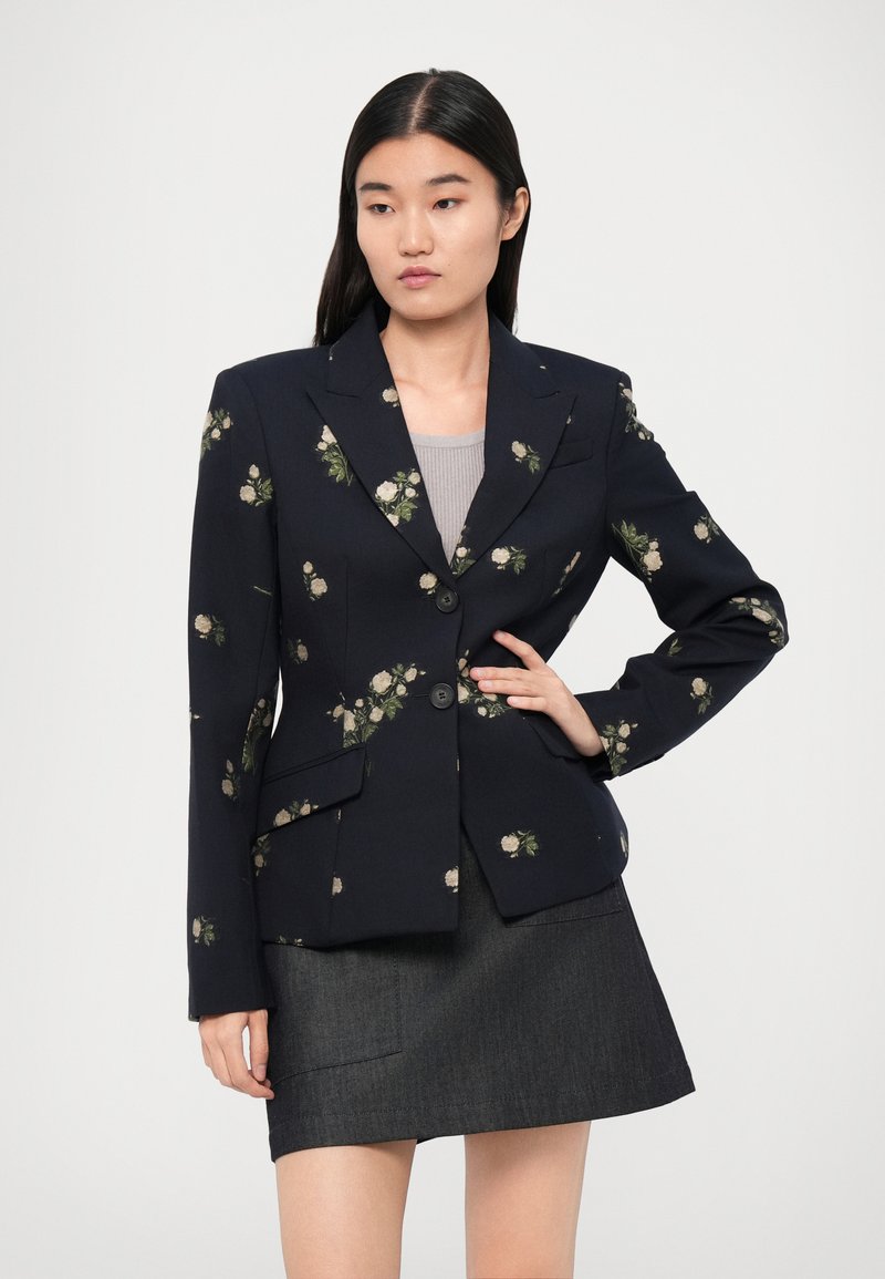 Marine blazer met bloemenpatroon en twee voorknopen. Beschikt over gestructureerde revers en twee voorzakken. Draagt over een grijze top en donkere rok.