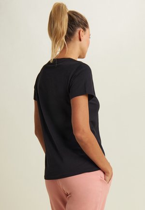 T-shirt nera in cotone con maniche corte e scollatura rotonda, abbinata a pantaloni della tuta rosa, mostrata di spalle. Texture morbida e vestibilità rilassata.