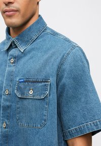 Kortärmad denimskjorta i ljusblått med knappad krage, framfickor med klaff och silverknappar. Har tydlig sömnad.