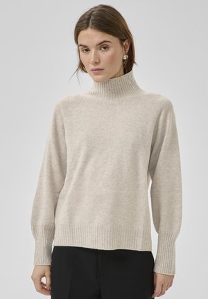 Donna che indossa un maglione a collo alto beige a coste con maniche lunghe e pantaloni neri, su uno sfondo chiaro uniforme.