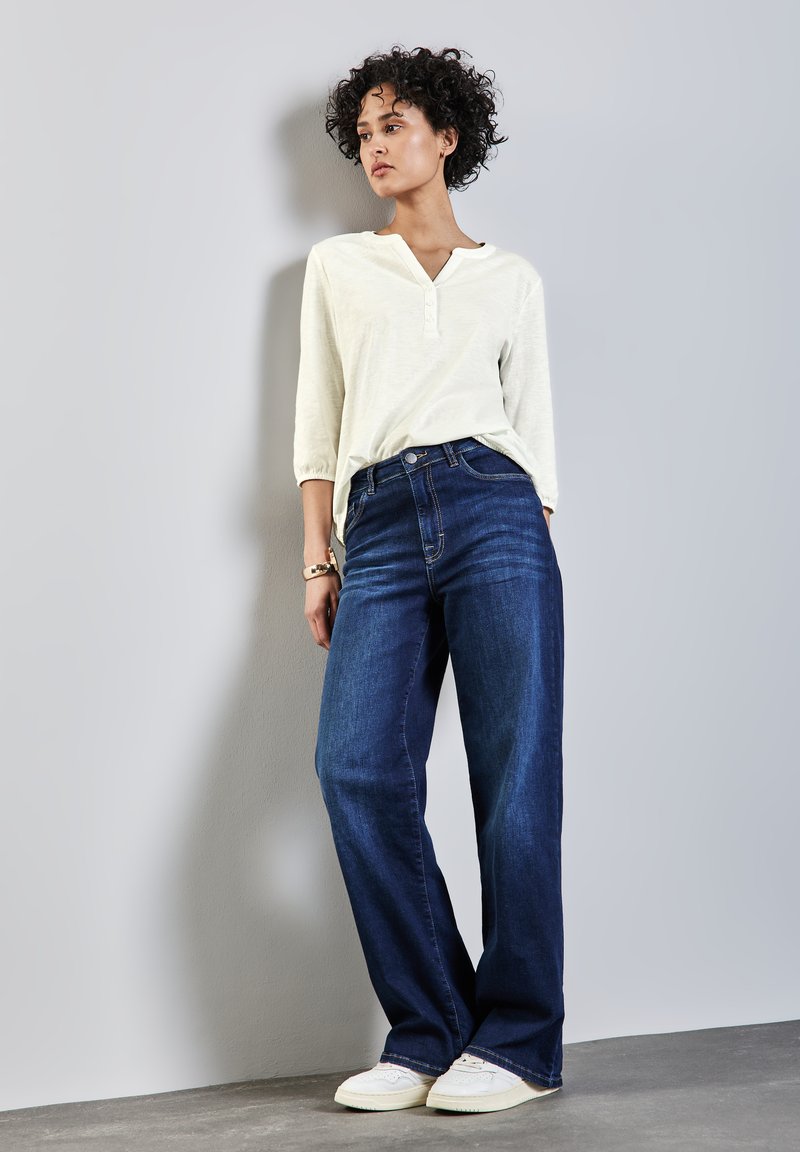 Maglietta henley beige chiaro con maniche a tre quarti, abbinata a jeans a vita alta blu scuro a gamba larga e sneakers bianche. Accessori minimi.