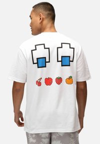 Camiseta blanca de algodón con un diseño gráfico en la parte posterior que presenta formas pixeladas azules y frutas en arte pixelado, incluyendo cerezas, manzana, fresa y naranja.