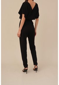 Mono negro con un diseño holgado de mangas cortas, un escote en V profundo en la espalda, cintura ajustada y piernas acampanadas, combinado con tacones negros. Textura suave.