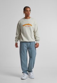 Sudadera gris claro con el texto "homeboy" y "est. 1988" en naranja y amarillo, combinada con pantalones de corduroy azul claro texturizados y zapatillas blancas.