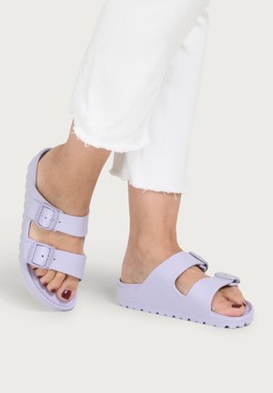 Feet wearing light purple open-toe sandals with two adjustable straps, paired with white cropped frayed hem pants.