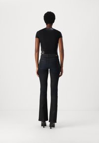 Nainen, jolla on lyhyet kiharat hiukset, seisoo selin päällään tiukka musta paita, korkeavyötäröiset tummat leveät farkut ja mustat korkokengät.