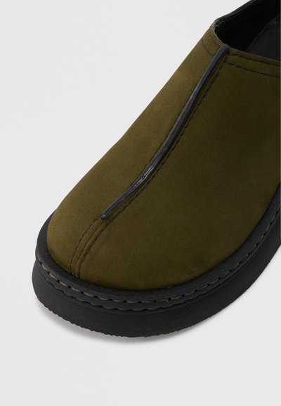 Παπούτσι slip-on από σουέντ ελιάς με στρογγυλεμένη μύτη, μαύρη καουτσούκ σόλα και μαύρες ραφές κατά μήκος των άκρων. Ομαλή υφή χωρίς σχέδια.