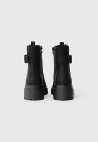 Bottes noires à la cheville en matériau texturé, dotées d'une fermeture éclair sur le côté, d'une accentuation de sangle, et d'une semelle en caoutchouc épaisse.