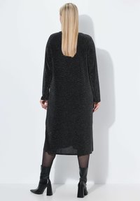 Langes schwarzes Kleid mit subtilen Glitzereffekten, langen Ärmeln, Seitenschlitzen und einer lockeren Passform. Getragen mit knielangen schwarzen Absatzstiefeln.