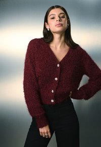 Vinrød, blød cardigan med v-hals, der har fem teksturerede knapper og ribbede manchetter. Kombineret med sorte bukser. Glat, blød tekstur.