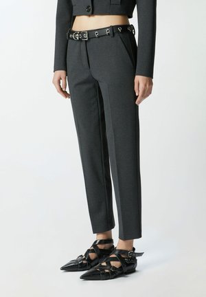 Pantaloni grigi su misura con una vestibilità cropped, dotati di pieghe frontali, tasche laterali e una cintura nera con accessori metallici. Ballerine nere a punta.