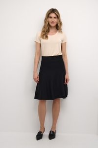 Haut beige à manches courtes avec un col rond associé à une jupe noire évasée jusqu'aux genoux. Le modèle porte des chaussures noires à bouts pointus.