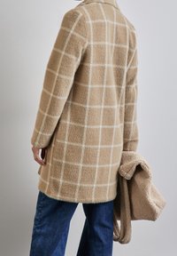 Abrigo de cuadros beige hecho de un material suave, con un patrón de cuadrícula blanca. Se sostiene en una mano una bufanda texturizada beige que acompaña.