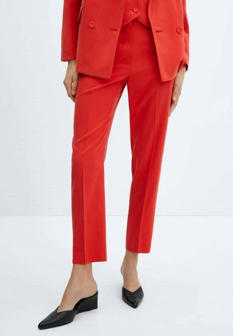 Blazer rouge ajusté et pantalons assortis, présentant un design structuré, un tissu lisse et une coupe slim. Des mules noires à bouts pointus complètent la tenue.