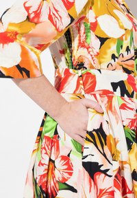 Blumenkleid mit großen rosa, roten und grünen Blumen auf einem cremefarbenen Hintergrund, mit einem gerafften Bund und seitlichen Taschen. Kurze Ärmel.