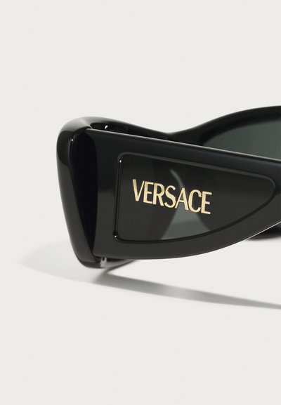 Melni saulesbrilles ar spīdīgu apdari, ar izteiktu zelta "VERSACE" logo sānos un tumšiem tonētiem objektīviem.