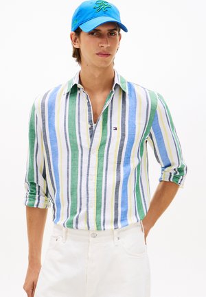 Junger Mann, der ein blau-grün-weiß-gelb gestreiftes Hemd mit aufgekrempelten Ärmeln, weiße Hosen und eine blaue Mütze trägt.