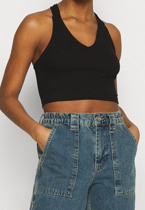Person iført en sort, ærmeløs cropped top og højttaljede blå denimjeans med hænderne i forlommerne.