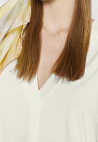 Haut blanc à col en V avec une texture lisse, associé à un foulard en soie jaune clair présentant un dégradé subtil. Cheveux longs et raides.